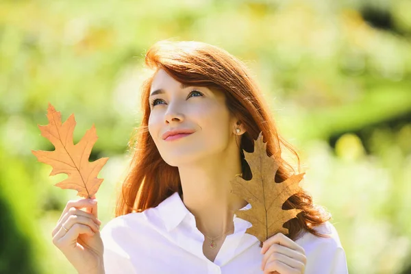 Romantic looking cute girl walking outdoor. Pretty woman playing with oak leaves. Woman with unbelievable nature beauty. Attractive dreamy girl with bright red hair and white blouse.