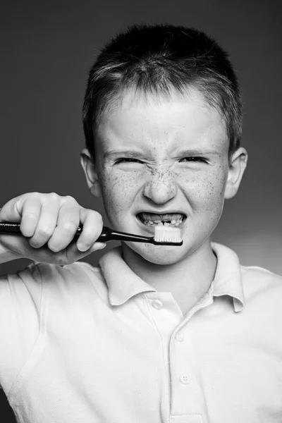 Red-haired boy with funny face is brushing teeth with toothbrush. Dental hygiene. Portrait of boy kid with toothbrush isolated on red background. Healthcare, dental hygiene, people and beauty concept. — Stock Photo, Image