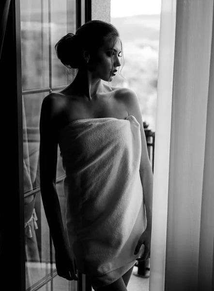 Αισθησιακό νεαρή γυναίκα τυλιγμένο σε πετσέτα μετά από ένα ντους στέκεται κοντά στο παράθυρο και κοιτάζοντας έξω. Όμορφη γυναίκα με πολυτελές μακιγιάζ και χτένισμα στο φόντο παράθυρο. — Φωτογραφία Αρχείου
