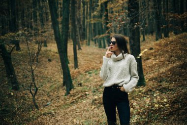 Woman walking at autumn nature in autumnal stylish outfit. Stylish femme fatale wearing fashionable clothes and smokes a cigarette at fall forest background.