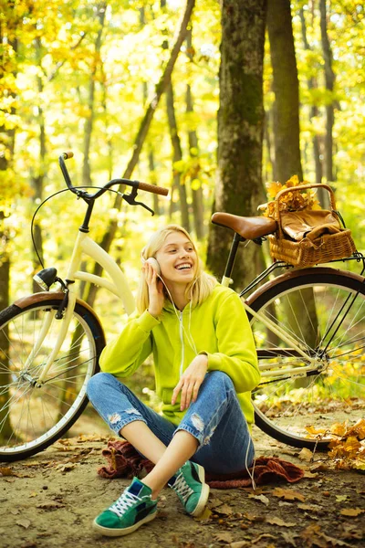 Vrolijk lachend tienermeisje zittend op herfstbladeren en luisterend naar muziek. Blond stijlvolle vrouw met fiets geniet van zonnige herfstdag in het bos. Actieve levensstijl en herfstseizoen concept. — Stockfoto