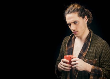 Handsome young man. In a bathrobe with a cup. Man with serious face and brutal style. Copy space. Side view portrait of confident man with beautiful hairstyle. Isolated on black background.