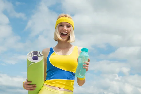 Concepto de agua e hidratación. Actividades deportivas al aire libre. Rubia alegre sonriente mujer sosteniendo deporte yoga estera y botella con agua. El agua es una necesidad vital durante los ejercicios deportivos . —  Fotos de Stock