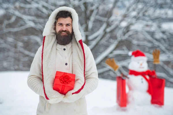 Buon Natale e felice anno nuovo. Divertente Babbo Natale uomo in posa con scatola regalo rossa sul tempo invernale. Pupazzo di neve e divertente uomo barbuto nella neve . — Foto Stock