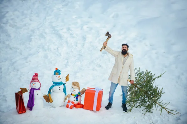 L'homme barbu porte un sapin de Noël dans le bois. Joyeux père avec sapin de Noël lors d'une promenade hivernale enneigée. Coupe-bois avec hache dans la forêt d'hiver . — Photo