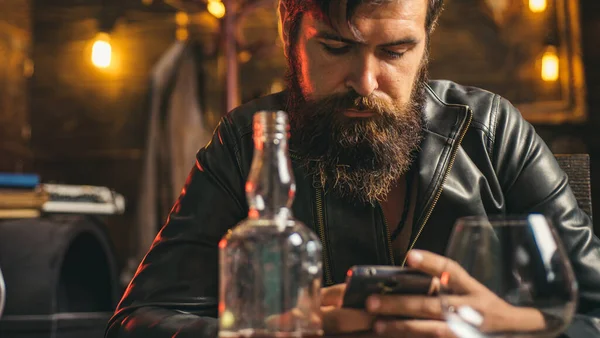 現代の実業家の生活様式。ハンサムな髭のビジネスマンは高価なウイスキーを飲んでいる。機械男は彼のスマートフォンでニュースフィードをスクロール. — ストック写真
