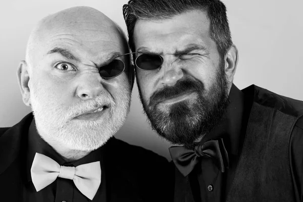 Father and bearded son are fooling around. Glasses for two. Men in suits and bow ties. Soul mates. Family business. Family traditions. Isolated on a light background.
