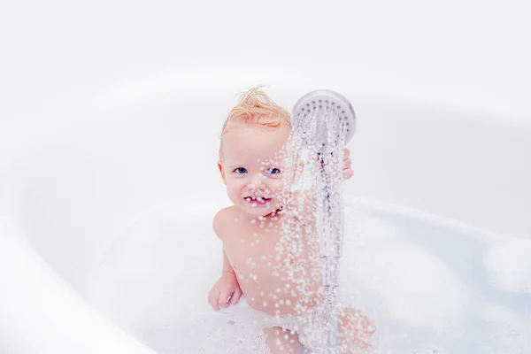 Улыбающийся мальчик принимает ванну с мылом. Милый маленький мальчик принимает ванну, играя с пеной и утиными игрушками в белой солнечной ванной. — стоковое фото