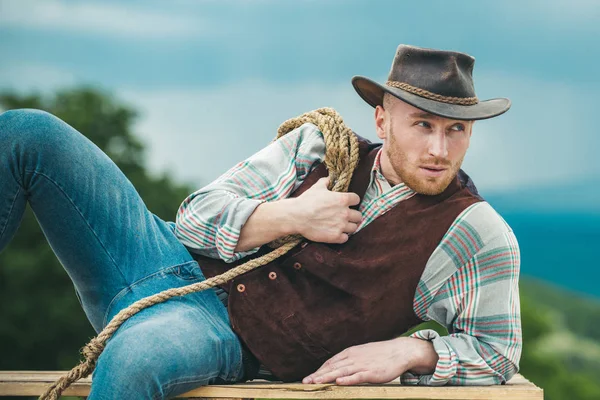 Sexy masculine stylish cowboy macho man holding lasso rope. Western style men fashion. Handsome cowboy in plaid shirt at beautiful rural nature background. — Stock Photo, Image