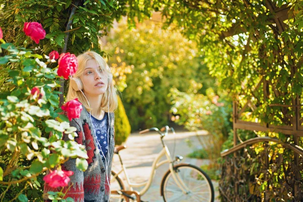 Hermosa mujer con estilo en traje de otoño disfrutar en el parque de otoño. Chica con auriculares se ve de ensueño. Rosas rojas y bicicleta al fondo. Clima cálido de septiembre. Concepto de otoño . —  Fotos de Stock