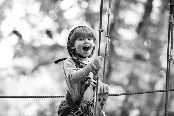 Cargo net climbing and hanging log. Portrait of a beautiful kid on a rope park among trees. Rope park - climbing center. Safe Climbing extreme sport with helmet.
