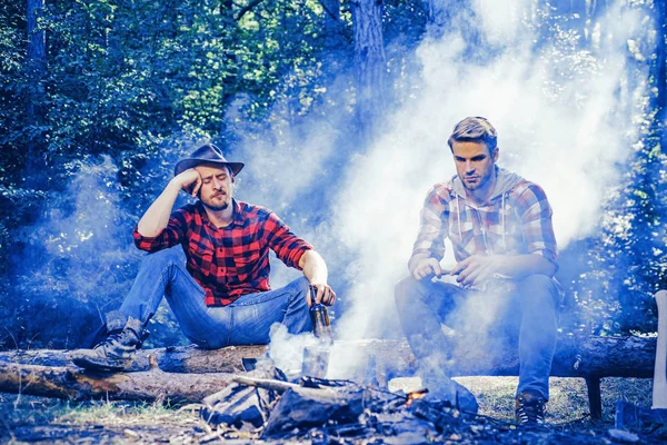 Two young guy sitting in the forest and drinking beer near campfire. Best friends spend leisure weekend hike barbecue forest nature background. Happy young male friends having picnic.