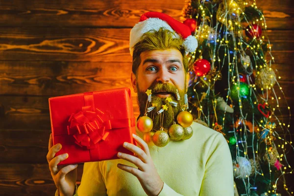Homme barbu s'amuser près de l'arbre de Noël à l'intérieur. Le Père Noël souhaite joyeux Noël. Père Noël dans le chapeau de Père Noël tenir cadeau de Noël . — Photo