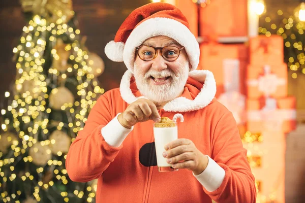 Święty Mikołaj z brodą i wąsami. Wesoły Święty Mikołaj trzyma szklankę z mlekiem i ciasteczkiem z kominkiem i choinką w tle. Święty Mikołaj w kapeluszu Santa. — Zdjęcie stockowe