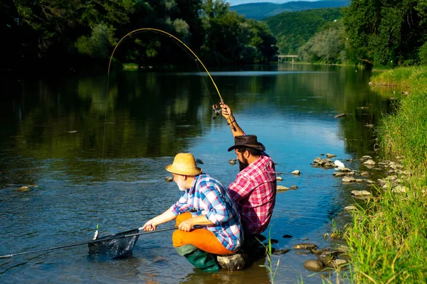 Pesca hobby maschile. Due amici maschi vestiti con camicie da pesca insieme a rete e canna durante la luce del mattino sul lago. L'uomo sul lungofiume godere di un paesaggio idilliaco e tranquillo durante la pesca . — Foto Stock
