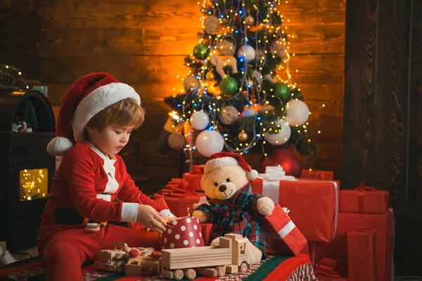 Le petit enfant porte des vêtements de Père Noël, avec un cadeau de Noël dans les mains. Le gamin attend le Nouvel An. Lumière de Noël dans une boîte cadeau . — Photo