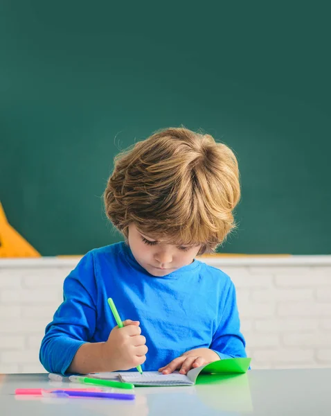 Glad söt flitig barn sitter vid ett skrivbord inomhus. Söt elev med roligt ansikte skolarbete. Barn gör sig redo för skolan. — Stockfoto