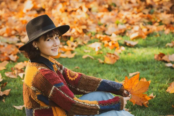 Regina d'autunno indossa maglione accogliente. Foresta autunnale nei colori oro e giallo. Donna hipster con cappello vintage che cammina nel parco. Affascinante bruna in cappello con foglie autunnali . — Foto Stock