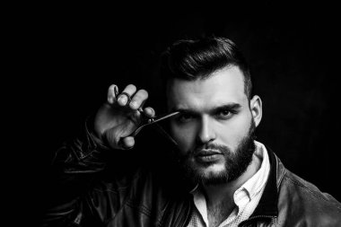 Bearded man, bearded male. Portrait of stylish man with beard. Barber scissors and straight razor, barbershop. Handsome barber is holding scissors near eye on black background. Stylish hairdresser.