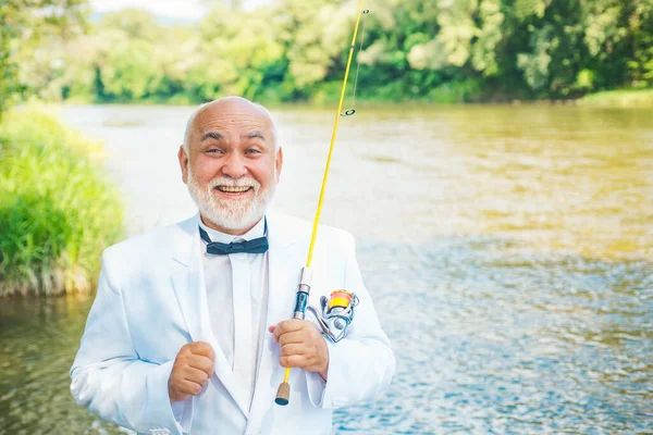 Retrato de un alegre hombre mayor pescando. Día soleado activo. Hombre pescando en el lago. Hombre barbudo pescando peces. Pescador barbudo en el agua. Pescador barbudo retirado. La leyenda se ha retirado . —  Fotos de Stock