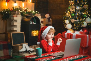 Noel çocuğu Noel arkaplanında Noel Baba 'ya evdeki bilgisayarda mektup yazıyor. Küçük çocuk dizüstü bilgisayarının yanında oturan Noel Baba kıyafetleri giyiyor. Not defteri kullanarak Noel Baba yardımcısı.