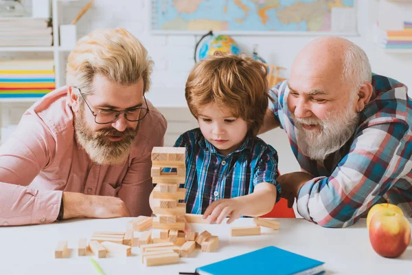 親の子供時代の値週末。おじいちゃんとお父さんが教室に座っていると、かわいい幸せな男の子がジェンガをしています。男性多世代の肖像画。家族のためのJengaゲーム. — ストック写真
