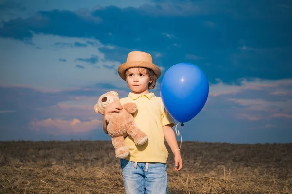 日没の背景におもちゃのテディと風船でフィールドに小さな男の子。庭の外で幸せな男の子。夏の野外で幸せな子供. — ストック写真