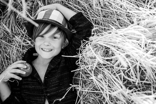 Boy in de hoed bedrijf apple en ligt op het hooi. Schattige kleine kind jongen houdt van bladgoud op boerderij dorp achtergrond. — Stockfoto