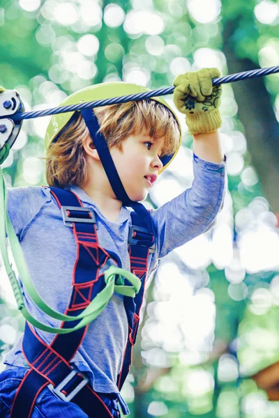 Cute child in climbing safety equipment in a tree house or in a rope park climbs the rope. Happy little child climbing on a rope playground outdoor. — Stock Photo, Image