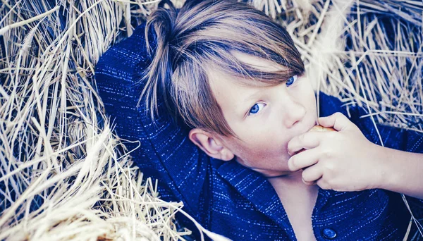 Divertido chico lindo se encuentra en el fondo del heno y se come una manzana. El chico anuncia ropa de niños para el otoño. Feliz infancia. . — Foto de Stock