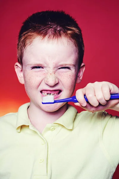 Рыжий мальчик со смешным лицом чистит зубы зубной щеткой. Гигиена зубов. Портрет мальчика с зубной щеткой изолирован на красном фоне. Здравоохранение, гигиена зубов, люди и концепция красоты . — стоковое фото