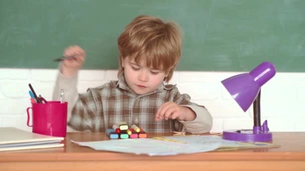 Zpátky do školy. Školáci kreslí proti zelené tabuli. Školák se šťastným výrazem na tváři u stolu se školními potřebami. Vtipný chlapeček ukazující na tabuli. — Stock video