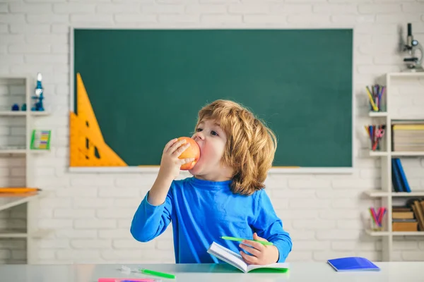 フレンドリーな子供は黒板の机の近くの教室でリンゴを食べる。小学校の教室。子供科学教育の概念. — ストック写真