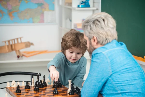 Concentrated little boy - father and son sitting at the table and playing chess. Teacher helping his pupil boy with chess on education class. Educational games.