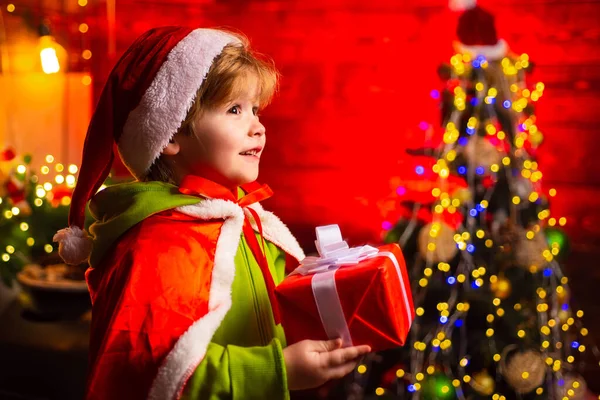 Glücklicher kleiner Junge am Weihnachtsbaum mit seinem Weihnachtsgeschenk, neugierig. Kleines Kind trägt Weihnachtsmannkleidung. Weihnachtskonzept. — Stockfoto