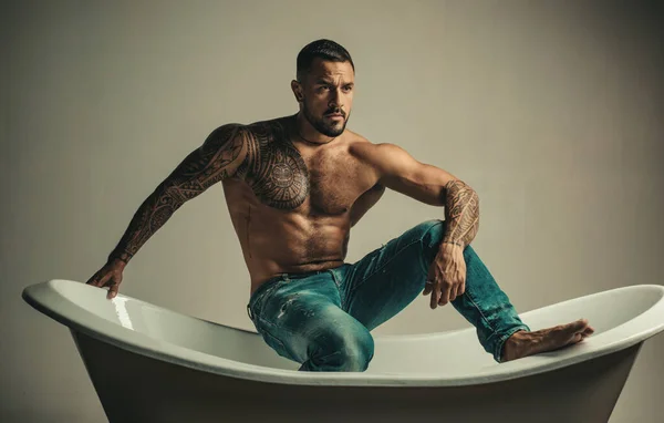 Homem machão sexual no banho. Homem tatuado musculoso forte segurando garrafa de champanhe e posando no banheiro. Bonito barbudo sem camisa homem em jeans com corpo sexy no banheiro . — Fotografia de Stock