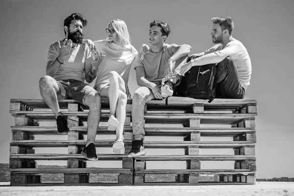 Charming young bearded man expressively tells his friends an interesting history. Company of friends students meeting after classes. Best friends sitting on bench at sky background.