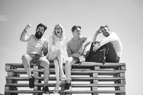 Company of friends students feel happy after passing the exams. Crazy bearded man and blonde woman. Group of four friends sitting on bench at clear sky background.