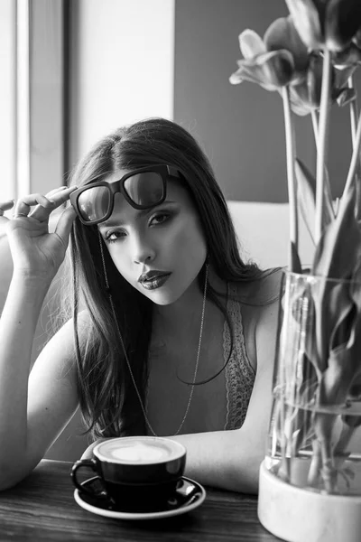 Een jonge vrouw die poseert in het café. Goedemorgen koffie. Een kopje. Een cappuccino. Bloemen op tafel. Goedemorgen vergadering. Sensueel model meisje met gladde huid en make-up. — Stockfoto