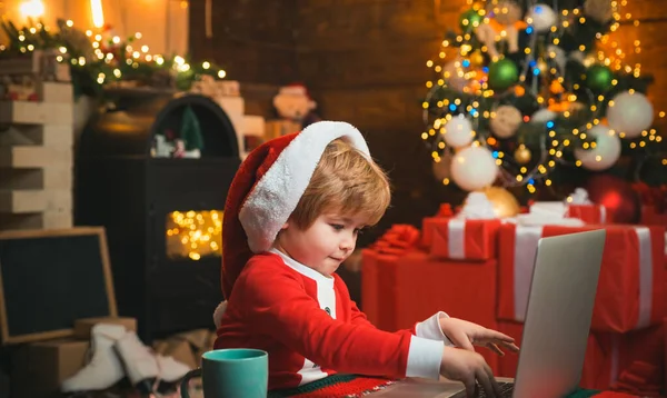 İnternetten çocuklar için Noel alışverişi. Mutlu, gülümseyen çocuk Noel Baba kıyafetleri giyiyor ve dizüstü bilgisayarına mektup yazıyor. Şömine arka planı. Tatillerde. Küçük çocuk Noel Baba kıyafeti giyiyor.. — Stok fotoğraf