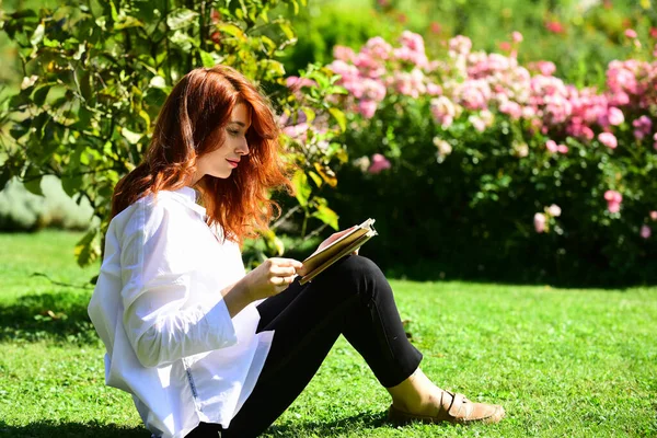 Kvinnlig student sitter i gräs med bok. Flickstudent njuter av litteratur inomhus. Rött huvud ung student. Ung kvinnlig poesi med långt rött hår funderar på att läsa spännande historia. — Stockfoto
