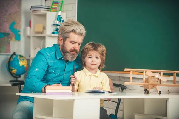 Kleiner Junge mit glücklichem Gesichtsausdruck neben Schreibtisch mit Schulmaterial. Nachhilfestelle. Lehrer zu Hause oder Hausaufgabenbetreuer. Vorschulbetreuung. — Stockfoto