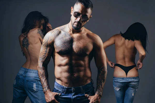 Sexy muscular macho man in sunglasses with two hot girls studio photo. Women with sexy fit ass standing behind handsome tattooed man with sexy torso. — Stockfoto