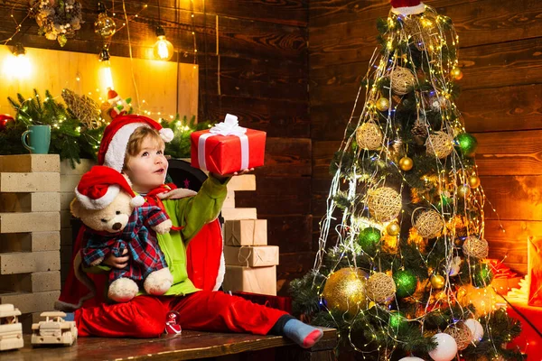 Счастливый маленький мальчик со своим плюшевым мишкой и рождественским подарком пытается угадать, что внутри подарка. Зима, отдых, семейное время . — стоковое фото