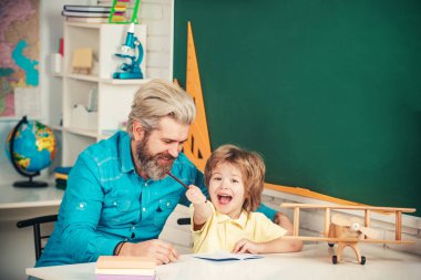 Baba oğluna öğretiyor. Ev ödevi. Okul malzemeleriyle masasının yanında gülen surat ifadesi olan küçük bir çocuk. Öğretmen ve öğrenci sınıfa. İlkokul sınıfı..