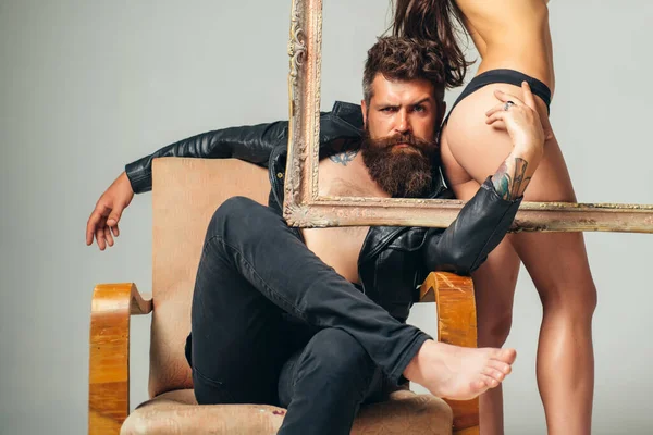 Bare hot girl with sexy ass standing behind a frame. Handsome muscular bearded man holding frame with sexy hot woman. Abstract art. Sexy couple isolated at grey background. — Stock Photo, Image