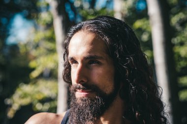 Ormanın arka planında dikilirken uzun siyah saçlı yakışıklı ve güçlü bir adamın portresi. Seksi sakallı, model maço adam. Erkek modası ve tarz kavramı.