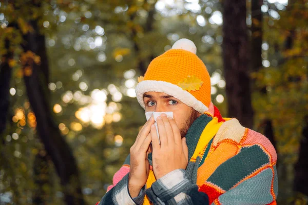 秋の公園でハンカチで男をくしゃみ.ナプキンでくしゃみをしようとしている病人。インフルエンザの流行を止めなさい。病気の男性は風邪を引く。少年は風邪をひき、紙のナプキンに鼻を吹く. — ストック写真