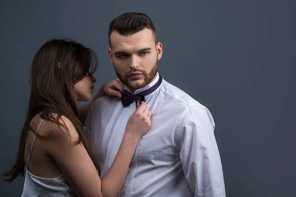 Ritratto di coppia elegante che posa insieme su sfondo grigio studio. Elegante accessorio maschile moderno. Donne che tengono il papillon pronto a vestirlo sull'uomo in camicia bianca. Coppia si prepara per l'evento . — Foto Stock