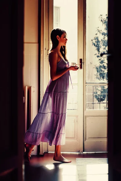 Вона тримає чашку, яку вона щасливо танцює. Дівчина одягнена в нічний одяг. Балкон з видом на будинок. Портрет красивої дівчини з довгим волоссям. Схід сонця вранці . — стокове фото